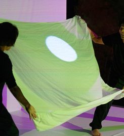 A Japanese man and woman are holding a sheet beween them. On the sheet a white circle of light is being projected. The man is smiling.