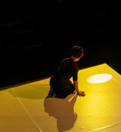 A woman is kneeling on stage, lit by a yellow light. Her back is to the camera, and she is looking at the floor where there is an oval of light being projected.