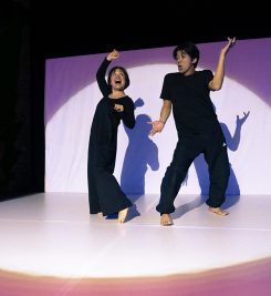 A Japanese man and woman, dressed in black, are dancing on a stage, lit by a giant white spotlight.