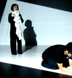 In the right hand corner of the photo, a japanese man wearing black kneels on the floor looking sad. To the left is a japanese woman standing, cuddling a white blanket partially covering her face. He has his back to her.