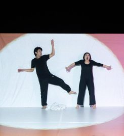 A Japanese man and woman, dressed in black, are standing in exaggerated positions up against a screen on a stage. They are lit by a giant white spotlight.
