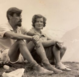 A black and white photo of a white man and white woman, both wearing shorts and t-shirts, sitting down. The woman is looking at the camera, but the man is looking into the distance. 